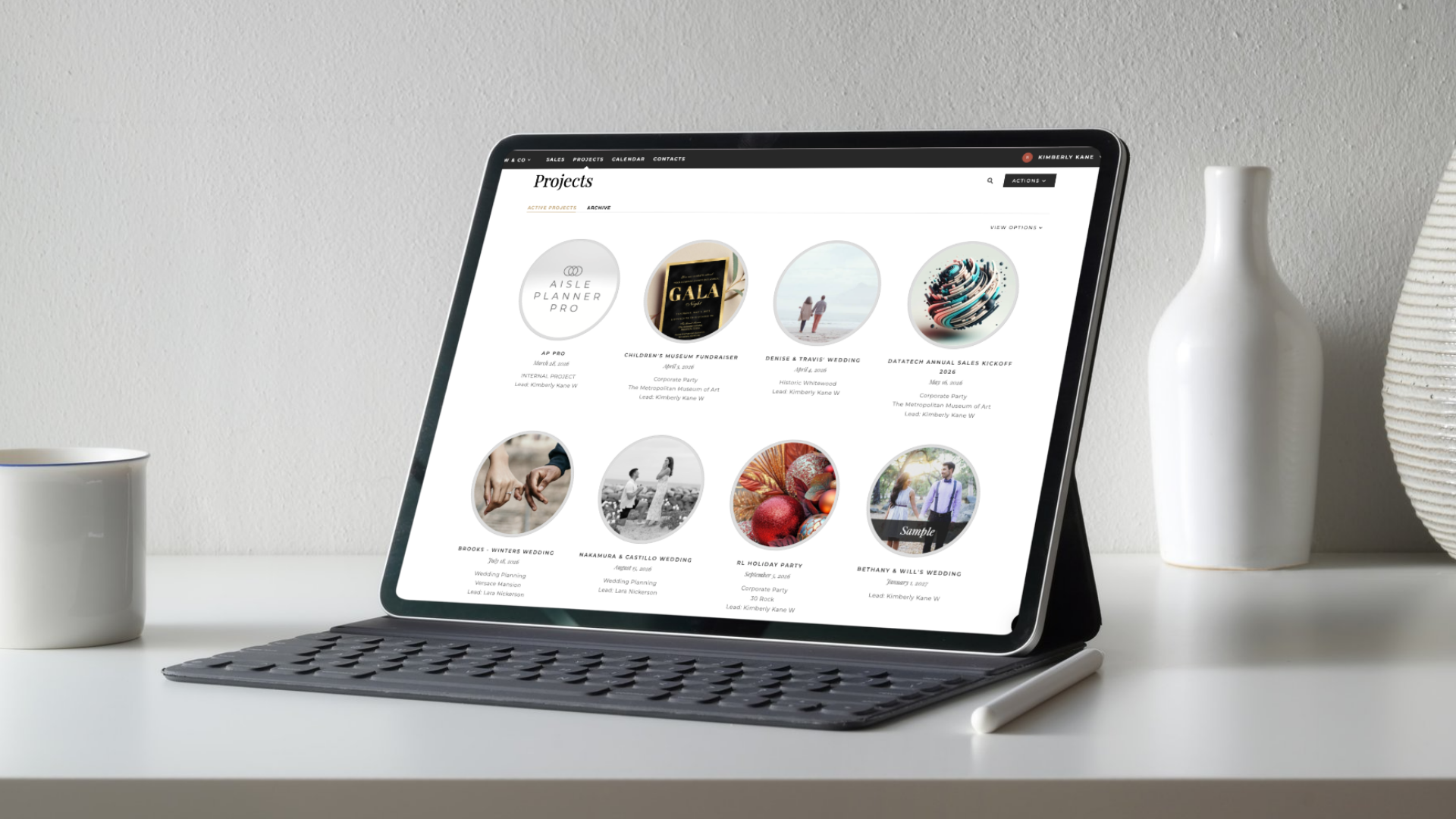 A professional tablet with a keyboard displays the Aisle Planner Pro wedding planning software and portfolio on a white desk, alongside a coffee mug and digital stylus for a modern event coordinator workspace.